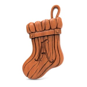 Op de afbeelding: Een bruine houten kerstsok-ornament met de letter "A". Het ornament heeft een getextureerd houtnerf-effect en een lus om op te hangen. Een decoratieve band omringt de bovenkant. Hoogte ongeveer 10 cm.