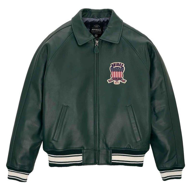 May include: Dark green leather jacket with a zip-up front, collar, and ribbed cuffs and hem with white stripes. Features a patch on the chest with the text "AVIREX" and a patriotic design.
