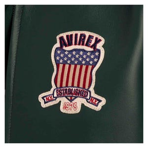 May include: A close-up of a dark green jacket with a patch. The patch has a shield design with red and white stripes, blue stars, and the text "AVIREX ESTABLISHED NY 1975". The patch is outlined in white.