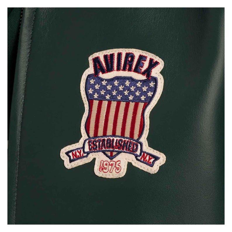 May include: A close-up of a dark green jacket with a patch. The patch has a shield design with red and white stripes, blue stars, and the text "AVIREX ESTABLISHED NY 1975". The patch is outlined in white.