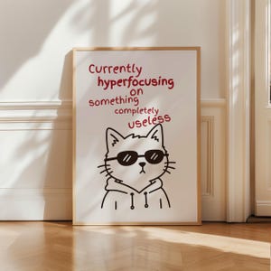 May include: A rectangular wall art print in a light wood frame featuring a black line drawing of a cat wearing sunglasses and a hoodie. Above the cat, red text reads: Currently hyperfocusing on something completely useless. The background is white.