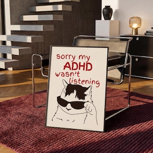 Può includere: Una stampa artistica rettangolare incorniciata con sfondo bianco e testo rosso che recita sorry my ADHD wasn't listening. Sotto il testo c'è un disegno a linee in bianco e nero di un gatto che indossa occhiali da sole scuri con una zampa alzata.