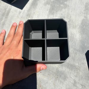 May include: A black, four-compartment organizer. The organizer is held by a hand and sits on a concrete surface. The compartments are square and the outer edges of the organizer have a geometric design.