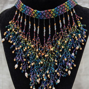 Op de afbeelding: Een veelkleurige kralenketting met een franje van kralen. De ketting is gemaakt van zwarte, blauwe, groene, gouden en witte kralen. De franje is gemaakt van zwarte, blauwe, groene, gouden en witte kralen. De ketting ligt op een zwarte fluwelen standaard.