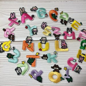 May include: A collection of embroidered alphabet patches in various colours, each with a different animal character. The letters are outlined in a contrasting colour, with details like flowers and fruit. The patches are scattered on a light wood surface.