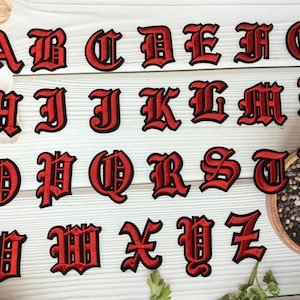 May include: A collection of red embroidered gothic-style alphabet letters, each with a black border. The letters are arranged on a light wood-grain surface, with some letters partially visible. The letters are in a bold, decorative font.