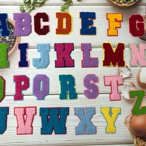 Op de afbeelding: Een verzameling kleurrijke, geborduurde varsity letter patches, elk met een andere kleur en textuur. De letters zijn gerangschikt op een wit houten oppervlak, met diverse voedingsmiddelen op de achtergrond.