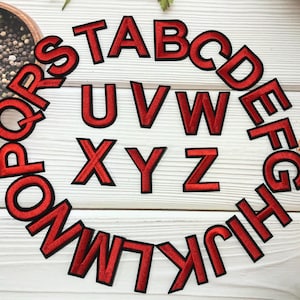 May include: Red embroidered alphabet letters with black outlines arranged in a circle. The letters are displayed on a white wooden surface. A bowl of black peppercorns is in the background.