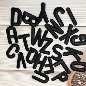 May include: A selection of black felt letters scattered on a light wood-grain surface. The letters are in various fonts and sizes, including the letters A, B, C, D, E, G, H, J, K, L, N, O, P, S, T, U, W, X, and Z. A small wooden bowl with peppercorns is in the background.