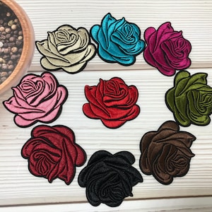 May include: A collection of embroidered rose patches in various colors, including cream, teal, fuchsia, pink, red, olive green, burgundy, black, and brown. The patches are arranged on a light wood-grain surface.