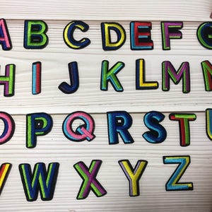 Puede incluir: Un conjunto de parches bordados con letras del alfabeto. Cada letra, con contorno negro, está rellena de colores vibrantes como rosa, verde, azul y amarillo. Los parches están dispuestos sobre una superficie de madera clara.