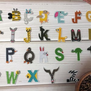 May include: A collection of colorful, embroidered alphabet letters, each designed with an animal theme. The letters are in various colors, including green, yellow, and brown, and are arranged on a light wood surface. The letters are suitable for children's crafts.