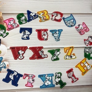 Puede incluir: Letras del alfabeto bordadas con personajes de Snoopy. Cada letra es de un color diferente, incluyendo rojo, azul, verde, amarillo y rosa. Las letras están dispuestas sobre una superficie de madera clara.