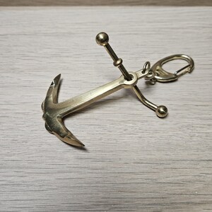 May include: A polished brass anchor keychain featuring a central shank with a crossbar and two curved flukes at the base. The top of the anchor is connected to a small metal jump ring and a spring-loaded lobster claw clasp for attachment.