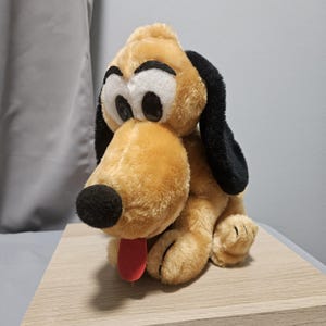 May include: A plush toy of a tan dog with long black ears, large white eyes with black pupils, black eyebrows, a round black nose, and a red tongue sticking out. The stuffed animal features soft fur and is depicted in a seated position.