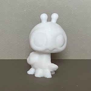 May include: A solid white figurine of a stylized creature with a large, rounded head, two prominent antennae on top, and oversized circular eyes. The figure has a smooth, matte surface and a compact body with small, rounded limbs.