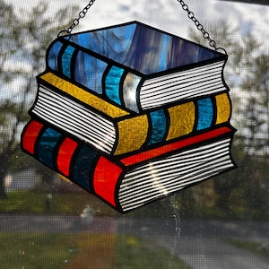 May include: A stained glass suncatcher shaped like a stack of three books. The top book is blue, the middle is yellow, and the bottom is red. Each book features textured white glass pages and dark blue vertical spine accents, all held by a metal chain.