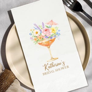 May include: White paper napkin with a watercolor illustration of a cocktail glass filled with flowers and a small pink umbrella. The text "Katherine's BRIDAL SHOWER" is printed in gold script.