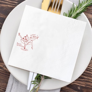 May include: White square paper napkin with a red illustration of a woman in a martini glass, with the text "A Tini Bit Older!" and "Emma's 30th Birthday". The napkin is on a white plate with gold silverware and rosemary sprigs.