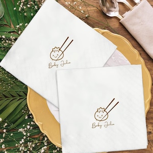 May include: White square napkins with a brown dumpling and chopstick design, and the text "Baby Julia". The napkins are placed on a gold-rimmed plate, with silverware and greenery in the background. Ideal for a baby shower.