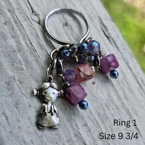 May include: A silver ring with a charm of a small girl and dangling purple, pink, and blue beads. The ring size is 9 3/4. The beads are various shapes and sizes, adding a playful touch to the jewelry.