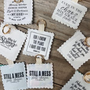 May include: A collection of square fabric keychains with scalloped edges, each featuring black printed text with religious quotes and phrases. Each keychain includes a beige fabric loop attached to a gold-toned metal ring for hanging.