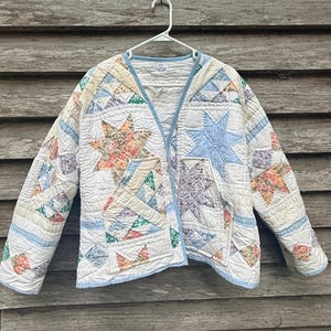 May include: A quilted jacket featuring a patchwork star pattern in shades of light blue, cream, and multi-colored floral prints. The garment has a V-neckline, long sleeves, two front patch pockets, and light blue trim along the edges.