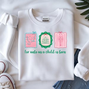 May include: White sweatshirt with three framed illustrations in pink, green, and blue. The illustrations depict a dove, a nativity scene, and a cross. The text "For unto us a child is born" is printed below the illustrations.
