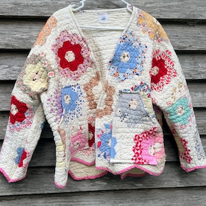 May include: A long-sleeve quilted jacket featuring a patchwork design of cream-colored fabric with various floral and geometric hexagonal patterns in red, blue, pink, yellow, and green. The jacket has a V-neckline and a pink scalloped trim edge.