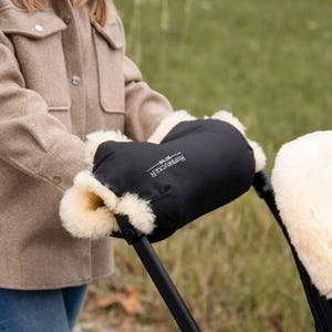May include: A black stroller hand muff with a cream-colored sheepskin lining, featuring the white text HOFBRUCKER SEIT 1948 printed on the front. The accessory is attached to the black handle of a stroller, providing warmth for hands.