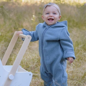 May include: A smiling baby wearing a blue hooded jumpsuit and a headband, holding a wooden toy stroller. The jumpsuit has a zipper down the front and is made of a soft, textured material. The stroller is white with natural wood handles.