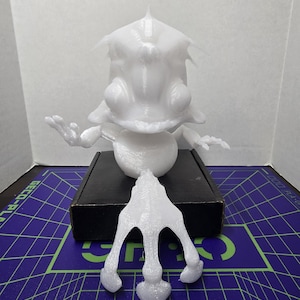 May include: A white 3D printed figurine of a creature with large eyes, a pointed head, and a wide mouth. The figure has a round torso, thin arms with three-fingered hands, and a separate, detached three-pronged base piece in front.
