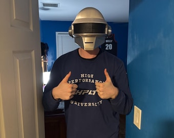 May include: A silver and black helmet with a rectangular visor, resembling a robot's head. The person is wearing a navy blue long-sleeve shirt with the text "HIGH PERFORMANCE HPLT UNIVERSITY". The person is also wearing black shorts.