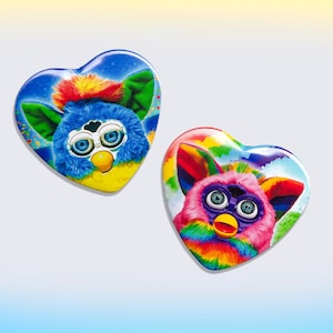 May include: Two heart-shaped Furby pins. One pin features a blue Furby with a red crest, green ears, and a yellow beak. The other pin has a pink Furby with rainbow accents. Both have the iconic Furby eyes.