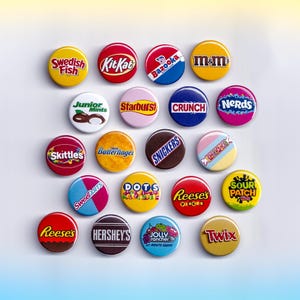 May include: A collection of colorful round pinback buttons featuring various candy brands, including Swedish Fish, Kit Kat, M&M's, Skittles, and Reese's. The buttons are arranged on a white surface.