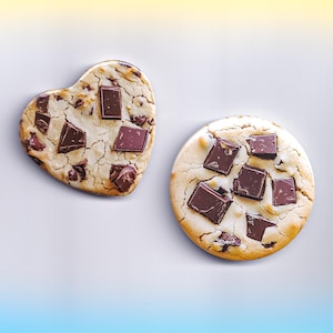 May include: Two chocolate chip cookies, one heart-shaped and one round, sit on a white surface. Both cookies have a light brown base with large, dark chocolate chunks. The heart-shaped cookie is on the left, and the round cookie is on the right.