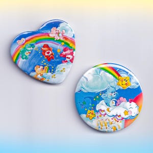 May include: Two Care Bears themed buttons. One is heart-shaped, the other is round. Both feature a blue sky background with rainbows, clouds, and Care Bears. The heart-shaped button has multiple Care Bears and a rainbow. The round button has a blue Care Bear on a cloud.