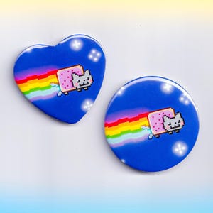May include: Two Nyan Cat-themed buttons. One is heart-shaped, the other is round. Both feature a pixelated cat with a Pop-Tart body, flying through a rainbow trail against a blue background. White star accents are scattered around the edges.