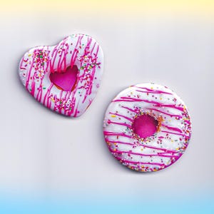 May include: Two glazed donuts, one heart-shaped and one round, with white frosting, pink drizzle, and colorful sprinkles. The heart-shaped donut has a pink heart cutout. A sweet treat.