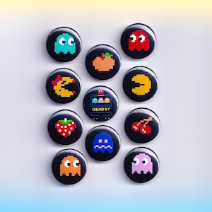 May include: A collection of black button pins featuring pixelated designs of video game characters and icons. The pins include a blue ghost, a red ghost, a peach, a crescent moon, Pac-Man, a strawberry, cherries, a blue ghost, an orange ghost, and a pink ghost. One pin reads "READY!"