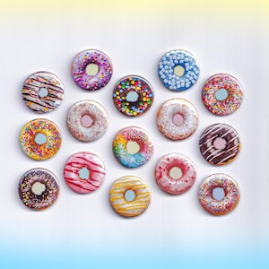 May include: A collection of colorful, round donut-shaped magnets with various toppings and designs. The magnets feature sprinkles, chocolate, frosting, and other decorative elements. The donuts are arranged on a white surface.