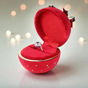 Op de afbeelding: Een rode fluwelen ringdoos in de vorm van een bol, open om een diamanten ring te onthullen. De doos heeft gouden accenten en een strik. De ring is gezet in een zilveren band.