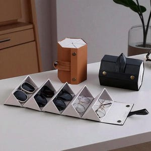 May include: Various eyeglass cases made of leather and fabric. A tan, hexagonal case with a handle, a black, rectangular case with a handle, and a white, triangular folding case are visible. The cases are designed to store glasses.