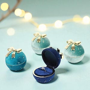 May include: A collection of velvet ring boxes in shades of blue and teal, each adorned with a gold bow. One box is open, revealing a diamond ring. The boxes are spherical and decorated with small white stars.