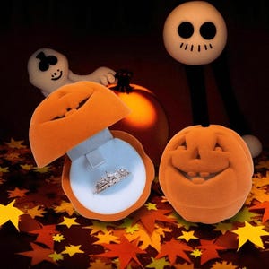 May include: An open orange pumpkin-shaped ring box with a silver ring inside. A second closed pumpkin-shaped box and a smiling ghost figure are in the background. A Jack Skellington-like figure stands behind the pumpkins. Autumn leaves are scattered on the surface.