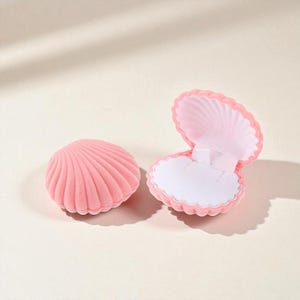 Puede incluir: Dos joyeros rosas con forma de concha. Uno está cerrado y el otro está abierto, revelando un interior blanco con un soporte para anillos. Las cajas tienen una textura suave y aterciopelada y están diseñadas para parecerse a una concha de vieira.