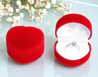 May include: A red heart-shaped ring box, open to reveal a silver ring with a clear gemstone. The box is velvet-textured and sits next to a closed matching box. White flowers are in the background.