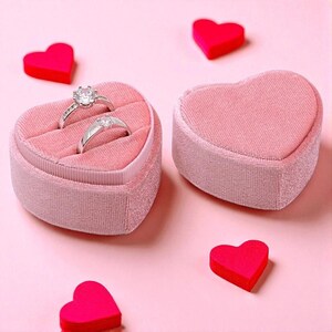 May include: Two silver rings with clear gemstones displayed in a pink heart-shaped velvet ring box. The box is open, revealing the rings nestled inside. Several red heart-shaped decorations are scattered around the box on a pink background.