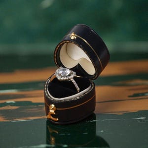 Puede incluir: Un anillo de compromiso de diamantes con una piedra central en forma de corazón se encuentra dentro de un joyero de cuero negro. El joyero está abierto, revelando el anillo sobre un interior de terciopelo negro. La caja está adornada con detalles dorados.