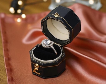 May include: Black, octagonal ring box with gold trim, open to reveal a heart-shaped diamond ring. The ring box is on a satin surface. The ring features a large, clear center stone and a delicate band.